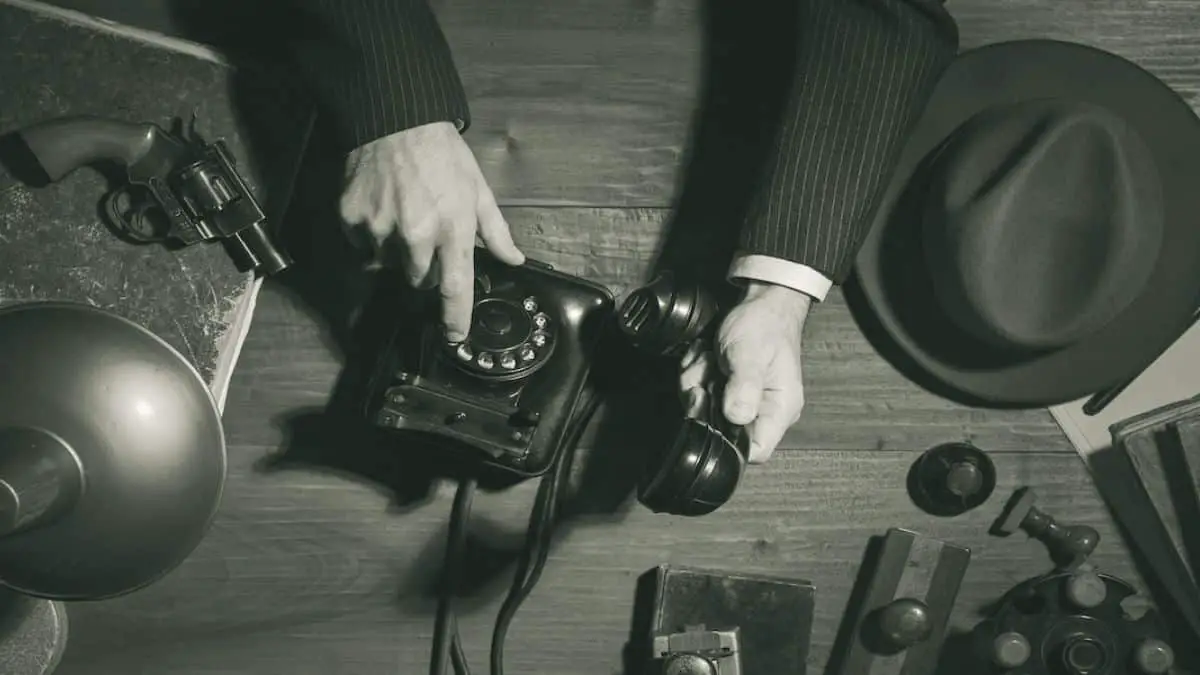 Man dialing vintage phone, revolver and hat on desk. (Difference Between Mystery and Suspense)